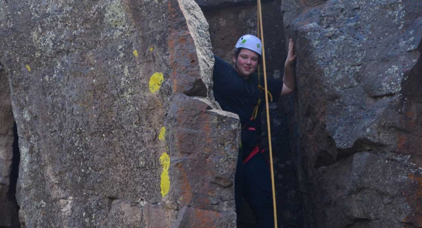 A student wearing safety gear smiles from a rock crevice while rock climbing.